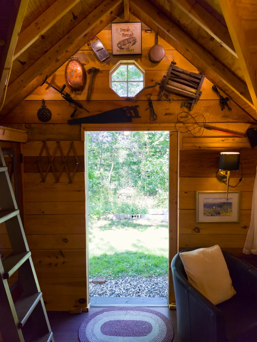 The cottage door open to the green woods beyond, vintage tools on the gable wall