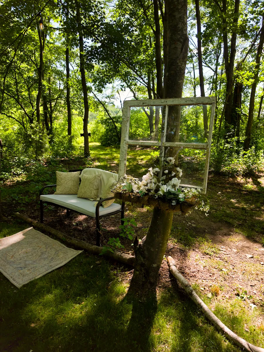 A cushioned bench beside a vintage window backdrop trailing white orchids in the mossy clearing
