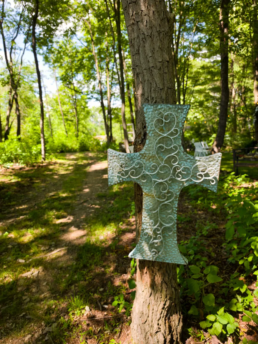 An ornate verdigris metal cross mounted on a tree along the wooded path into the chapel