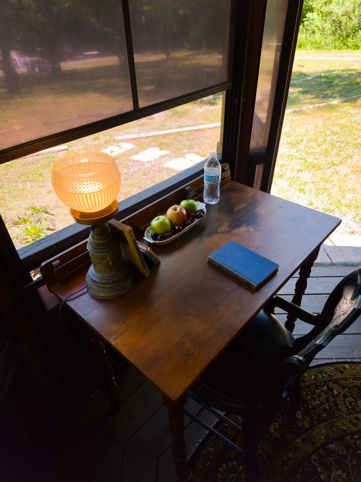 An antique writing desk by the screened window with a lamp, books, and a bowl of fruit