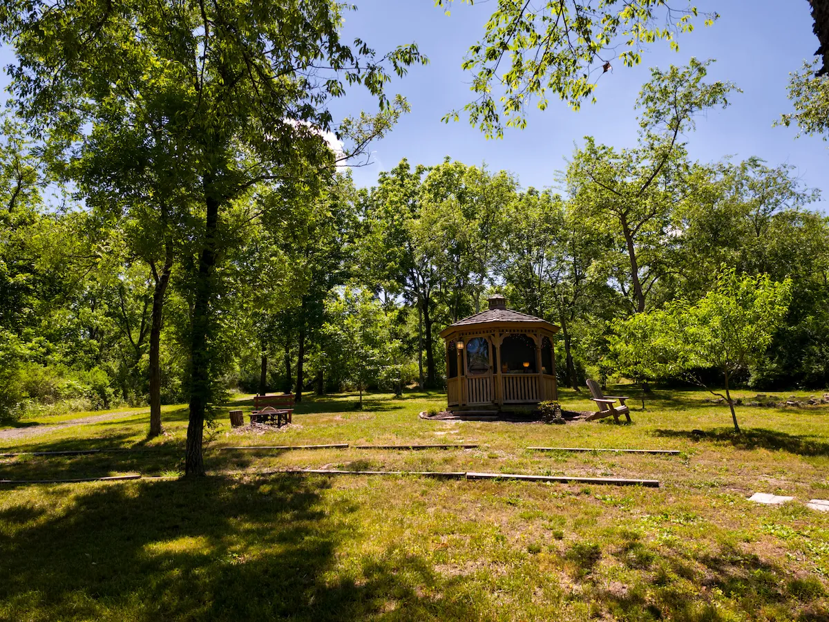 Ground-level aerial of the gazebo nestled in the wooded field with a fire pit and Adirondack chairs nearby