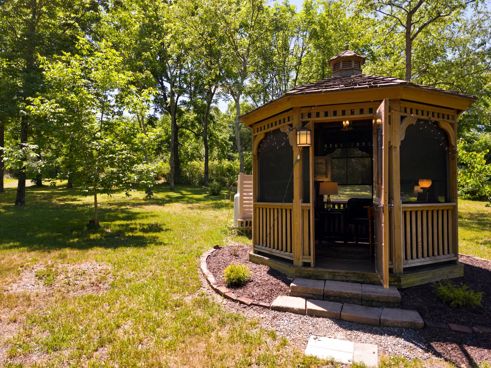 Wide interior of the gazebo at dusk, lit from within, set among the trees in the open field