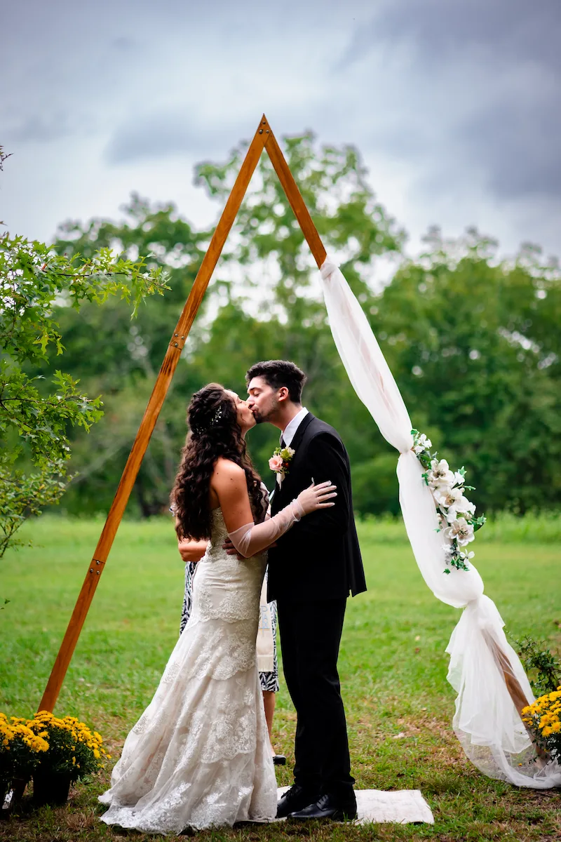 Alex and Naomi kiss beneath the wooden A-frame arch in the open field